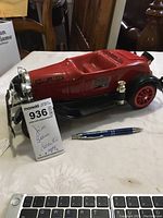 Full side view of red fire department themed Jim Beam decanter shaped like a vintage fire truck with chrome details and black base, placed on a table next to a pen for scale and the auction tag.