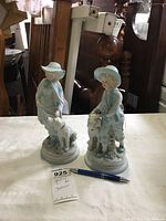 Photo showing two blue and white bisque statues of girls with dogs on a white surface. Background items not included.