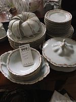 Photo showing stacked mismatched Heinrich Duchess china plates, cups hanging on edge of plates, and a lidded serving dish on wooden table.