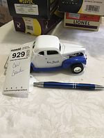 Side view of metal model car key bank with MaxSold tag showing lot number 929 and note 'Car Bank' alongside pen for scale.