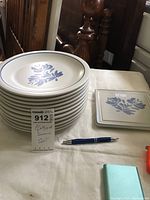 Stack of eleven Pfaltzgraff dinner plates beside a matching trivet, pen for scale, on a table.