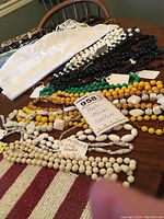 Photo showing various plastic beaded necklaces in multiple colors arranged on a wooden surface. Necklaces include white, black, yellow, green beads with some oblong and twisted bead designs.