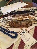 Collection of seed bead necklaces laid on a white cloth background showing various colors and styles including black seed beads, gold tubular beads, blue twisted beads and brown round beads.