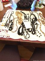 Photo of a variety of sparkly beaded necklaces arranged on a white cloth on a table. The necklaces include mostly black beads, some goldtone beads, a hematite necklace, and pearl-colored beads.