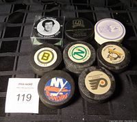 Photo showing seven hockey pucks with various team decals including Jean Ratelle Team Canada 1972 commemorative puck, Boston Bruins, New York Islanders, Philadelphia Flyers, Pittsburgh Penguins, Vancouver Canucks, and a white puck with purple logo.