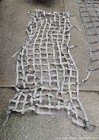 Grey nylon strap cargo net about 6'x3' with metal rings at corners laid flat on concrete.