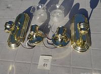 Four brass wall light fixtures with brass backplates laid out on surface with two frosted glass shades positioned above.