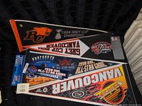 Four sports pennants laid flat: two BC Lions Grey Cup pennants, one Vancouver Canucks NHL All-Star Game 1998 pennant with stadium image, and one Vancouver Canucks colorful logo pennant.