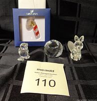 Four Swarovski crystal ornaments arranged on black fabric including crystal duck, fox, faceted ball, and boxed candy cane ornament