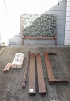 Photo showing all disassembled wooden bed frame components laid out on a concrete surface, including footboard, side rails, and slats along with the padded headboard.