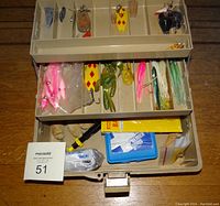 Top view of tackle box with open tray showing assortment of fishing lures and soft plastic baits in sections.