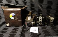 Three cameras placed horizontally with Kodak carrying case on left, Polaroid Minute Maker in center, two Kodak Brownie Hawkeye cameras on right.