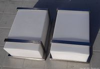 Two rectangular chrome and frosted glass wall light fixtures placed on tiles under sunlight, showing the front and top views.