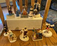 Overall view of the 10 miniature figurines displayed on a wooden surface and a raised white box, showing a variety of historical and themed figures in period clothing.