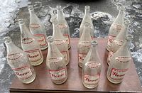 Twelve clear vintage soda bottles displayed standing upright on wood, showing intact fire-glazed red graphics with brand and location text.