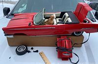Red vintage remote control toy car with open trunk displaying spare tire, detachable front tire, remote controller with wire, and seated driver figure.