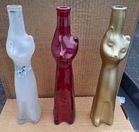 Three cat-shaped glass wine bottles of gold, red, and frosted clear glass standing side by side on cardboard, showing their overall shapes and user-visible condition.