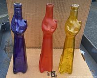 Full view of the three cat-shaped colored wine bottles in deep blue, orange, and yellow, standing on a cardboard backdrop outdoors.