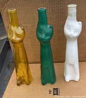 Three tall cat face wine bottles standing upright against cardboard background showing white, greenish, and yellow bottles.