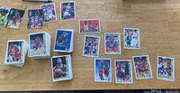 Wide shot of multiple basketball cards spread on a wooden surface, showing a mix of cards and stacks, including various NBA players in action poses.