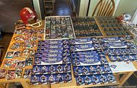 Full view of the lot showing multiple uncut sheets of football cards spread on a table along with a vintage red full-size football helmet and a white mini New England Patriots signed helmet.