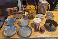 Wide shot of various tins, piecrust pans, Red Flash Cleaner tins, Uncle Ben's rice tin, tennis ball container, and picnic meal warmer spaced on a table.