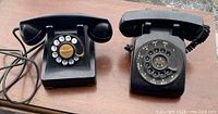 Two black rotary telephones shown side by side on a wooden surface, one vintage with old cord and one modern with wall connection cord
