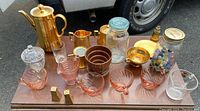 Photo showing entire lot on wooden table with a vehicle tire in background, items include gold coffee pot, pink glass dishes, jars with marbles, salt and pepper shakers, and other vintage glass and ceramic pieces.