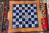Top-down photo of the chessboard with all chess pieces arranged on the sides inside two red velvet lined trays. Board has decorative painted white squares and alternating black squares, with one black square missing.