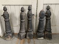 Five antique black cast iron fence posts lined up, showing full height and acorn finials on four posts.