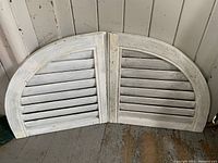 Pair of antique curved transom window shutters with white paint and visible wear, laid open against a gray wooden wall and floor.
