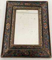 Photo shows front view of rectangular mirror with wide decorative handpainted tin frame. The frame has blue and burnt orange floral and oval medallion motifs with deliberate wear marks evident.