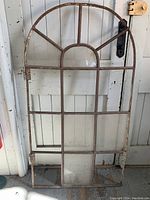 Full view of antique iron frame arched window leaning against a door, featuring a rounded top with multiple glass panes, some missing.