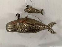 Two silvered glass whale ornaments on a white surface; larger about 6 inches, smaller above it.