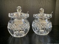 Two vintage Waterford crystal jam jars with lids and spoons standing side by side on a dark surface.