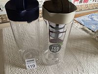 Two clear Komax pitchers, one labeled as a tea pot with infuser, both with dark lids, placed on a textured white cloth surface.