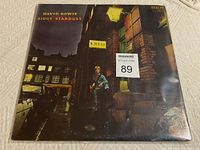 Photo showing front of David Bowie Ziggy Stardust album cover with street scene and Bowie in costume, some wear and creases on edges.