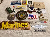 Photo showing an assortment of military items including patches, pins, stickers, postcards, and small figurines arranged on a beige surface.