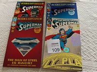 Four Superman comic books arranged in two pairs. Left pair shows two copies of 'Superman The Man of Steel' comic with a red cover and Superman emblem. Right pair shows two copies of 'The Adventures of Superman' comic with holographic silver cover.