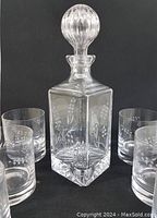 Clear hand cut crystal decanter with round ribbed stopper and etched leaf and berry design, surrounded by four whiskey glasses with matching etched pattern on a black background.