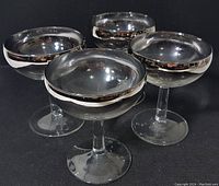 Four clear glass champagne coupes with silver fade rims shown in group on black surface. Each glass is approx 4.5 inches tall, 3.5 inch diameter at top. Vintage mid-century style with short stem and wide bowl.