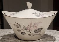 Full front view of bowl with lid on, showing oval shape, floral decoration and built-in handles on sides.