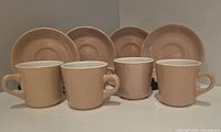 Set of four beige vitrified cups with matching saucers displayed in a slightly staggered row on white background.