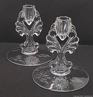 Two clear Hughes Cornflower glass candlestick holders on a black background. Both show floral and leaf embossed patterns on the round base, measuring about 5 1/8 inches diameter and 5 7/8 inches tall.