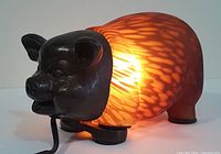 Pig-shaped lamp illuminated, showing glowing orange mottled art glass body with dark metal head and legs.
