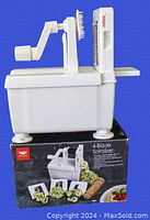 Side angle of white PADERNO 4-blade vegetable slicer and spiralizer mounted on its original box. Shows storage container base, crank handle, folding arm, blade holder, and suction feet.
