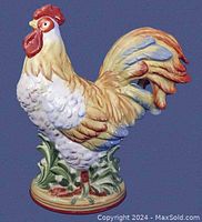 Full view of the hand painted ceramic rooster figurine with detailed paint and textured feathers, showcasing the entire piece from the side at knee height against a blue background.
