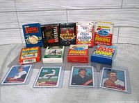 A front view of 11 boxed baseball card packs from brands Topps, Donruss, Tombstone, Kmart, Kraft, Leaf showing various editions and series from 1980s-1990s with 4 loose baseball cards laid out in front.
