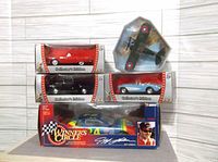 Photo showing five boxed diecast models including three Road Signature 1:43 cars, one Winner's Circle 1:24 NASCAR car, and one Edison Air line biplane in display case.