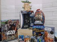 Photo showing Grogu plush, Darth Vader electronic figure with gumballs, and Force Battlers Yoda figure packaging alongside Star Wars books and playing card decks.